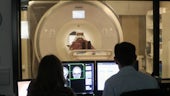 A participant lies in an MRI scanner while a male and female researcher operate the scanner