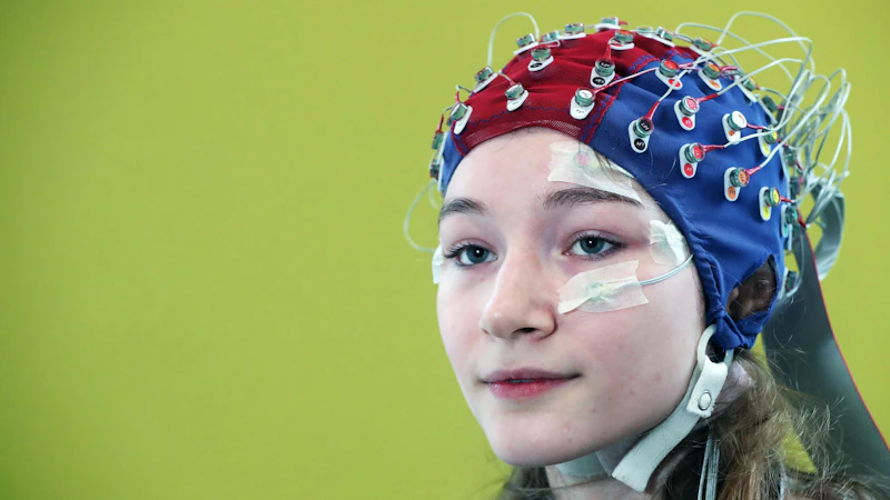 a female participant has a blue and red electrode cap on her head with numerous electrodes and wires attached for brain imaging