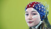 a female participant has a blue and red electrode cap on her head with numerous electrodes and wires attached for brain imaging