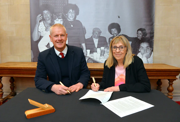 A man and a woman look at the camera as they sign a document on the table in front of them