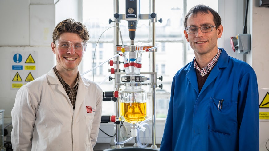 Two men wearing lab coats and protective eyewear are photographed in front of a chemical reactor in a lab at Cardiff University’s School of Chemistry.