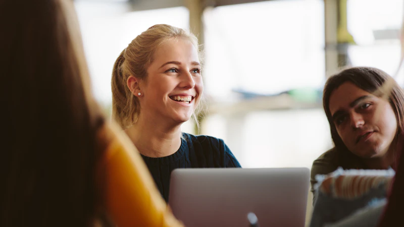 Three people sitting at a desk with their laptops open, the main focus is on a female with blonde hair who's smiling