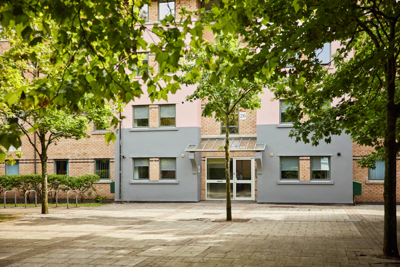 Entrance of a brick and gray apartment building, partly shaded by green trees
