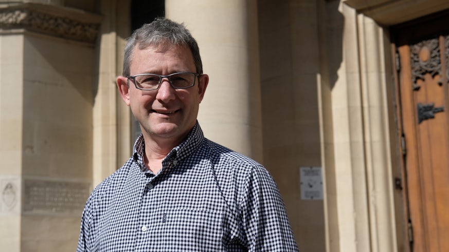 A man wearing glasses and a checked shirt is photographed outside a period building.