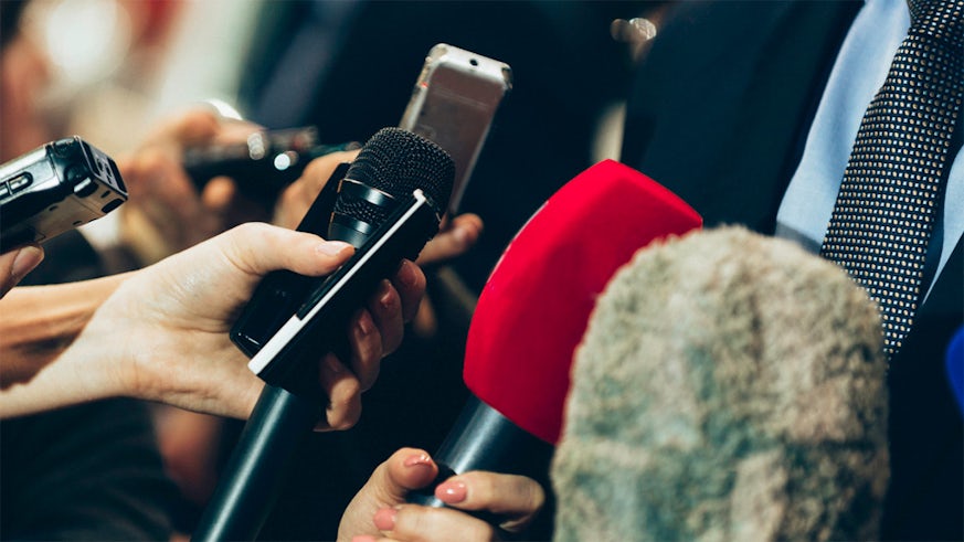 A man wearing a suit is interviewed by reporters holding microphones