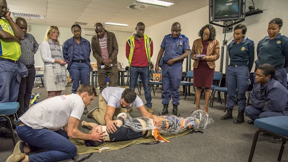 Two trainers kneeling on floor with a dummy model, watched attentively by police officers and others being trained