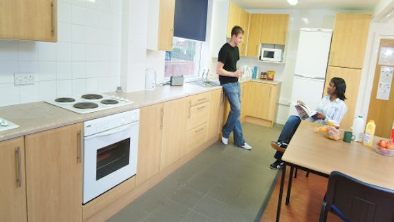 Kitchen area with two people talking, one seated, the other leaning against cabinet.