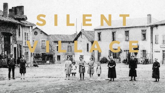 Black and white image of ten people (men, women and children) standing in a row, in the middle of a village, and facing the camera straight ahead.