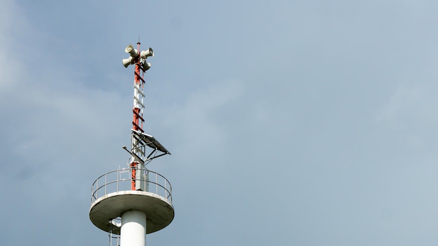 The top of a tsunami warning tower is photographed against a blue sky. The tower is painted red and white and features solar panels and megaphone speakers