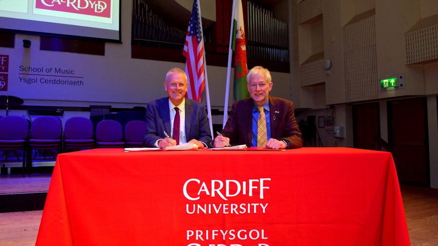 Two men wearing suits are sat at table draped with a cloth bearing Cardiff University’s logo