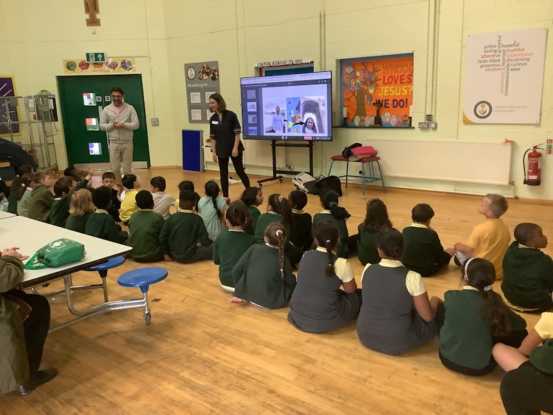 School children photographed in a classroom where adults are presenting on a whiteboard.