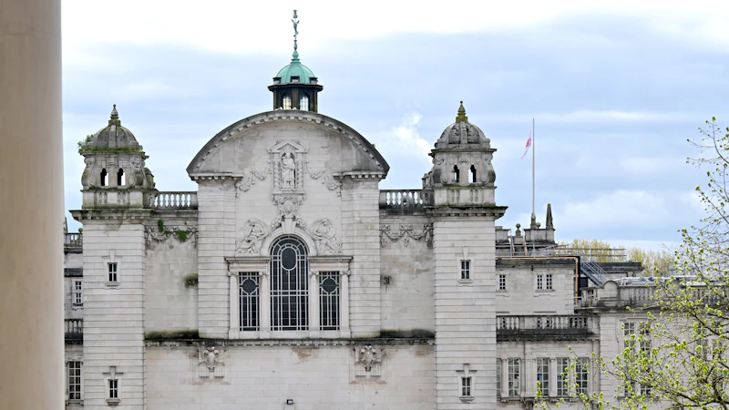 Front view of a historic university building with ornate stone architecture, large arched window, decorative carvings, and two small domed towers. A green copper cupola sits at the top center, and a flagpole with a flag is visible to the right. Trees frame the scene under a partly cloudy sky.