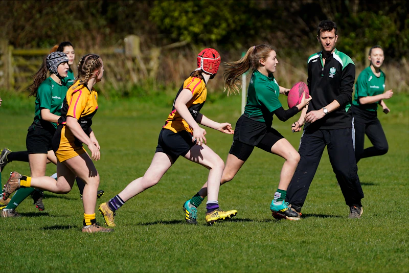 Teenage girls playing rugby with a pink ball. The teams' kits are green and black and yellow and black.