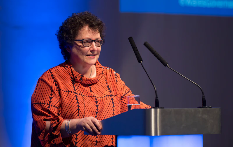 Elin Jones AM wears an orange top and speaks at a podium. 