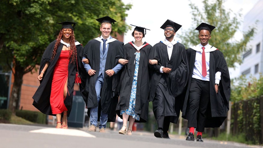 5 graduates in graduation gowns walking towards the camera