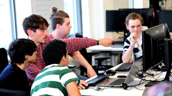 Image of students crowded around a laptop and monitor, discussing a project they are working on