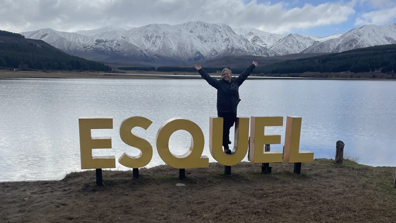A photo of a female standing behind a sign that says 'Esquel'. Behind the female there is a lake and mountains.