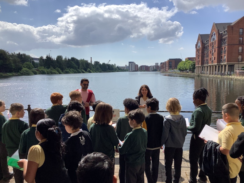 Two adults stand in front of a group of schoolchildren at an inner-city canal.