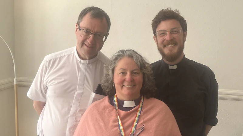 Three individuals wearing clerical collars stand together indoors, smiling. The person on the left wears a white shirt and glasses, the middle person wears a pink garment over a black shirt with a rainbow lanyard, and the person on the right wears a black shirt and glasses.