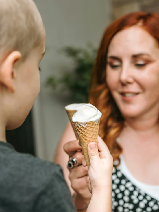A young woman with long red hair and wearing a black and white spotty top is facing a fair-haired male child, both are raising their ice cream cones to each other. There is vanilla ice cream in the cones.
