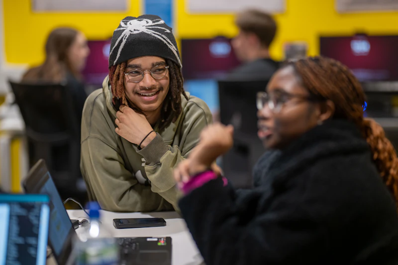 A photo of a student who is sitting at a desk and is smiling. The student is wearing a green top, a hat and glasses.