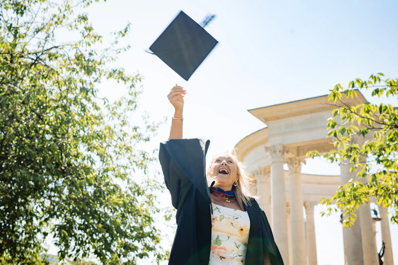A woman in graduation attire smiling and throwing a cap on a sunny day. 