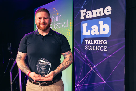 A man standing on stage at an event, talking to the audience with hand gestures. A large screen is in the background that reads 'Fame Lab Talking Science'. 