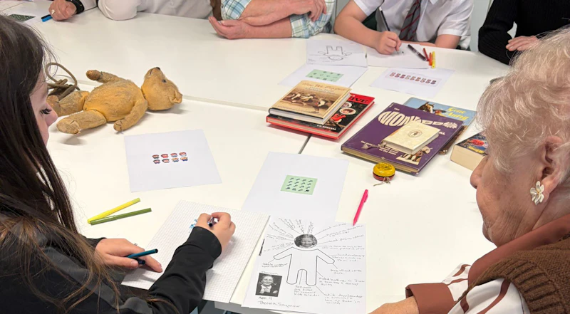 An older person and a teenager are facing away from the camera lens, working on designs on a piece of paper on a table. 