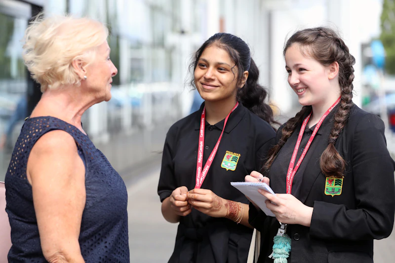 An older woman is interviewed by two female high school students dressed in black blazers. 