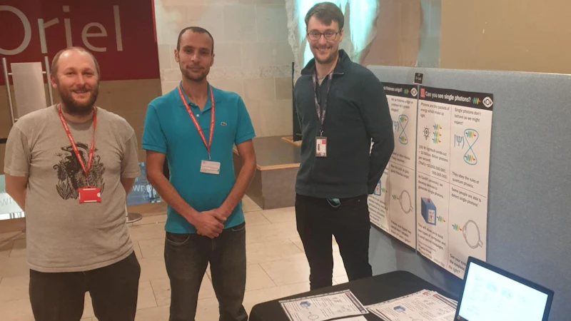 Three men pose for photograph in exhibition space. A table beside them has documents and a laptop on it. Posters with text and diagrams are displayed on a banner behind them.