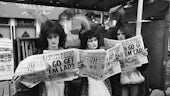 Three people in heavy makeup and wigs read Sun newspaper with Go Get Em Lads on the front pge in front of dramatic shopfront with wigs
