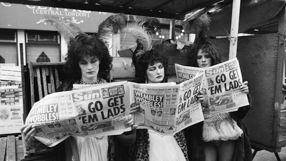 Three people in heavy makeup and wigs read Sun newspaper with Go Get Em Lads on the front pge in front of dramatic shopfront with wigs
