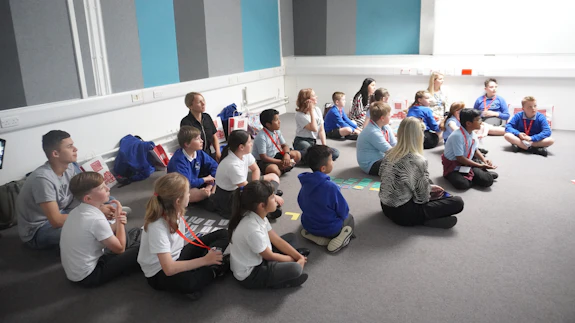 A group of boys and girls who are wearing school uniform are sitting on the floor.