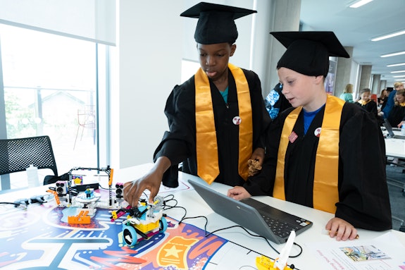 Two children in caps and gowns look at a laptop and toy models.
