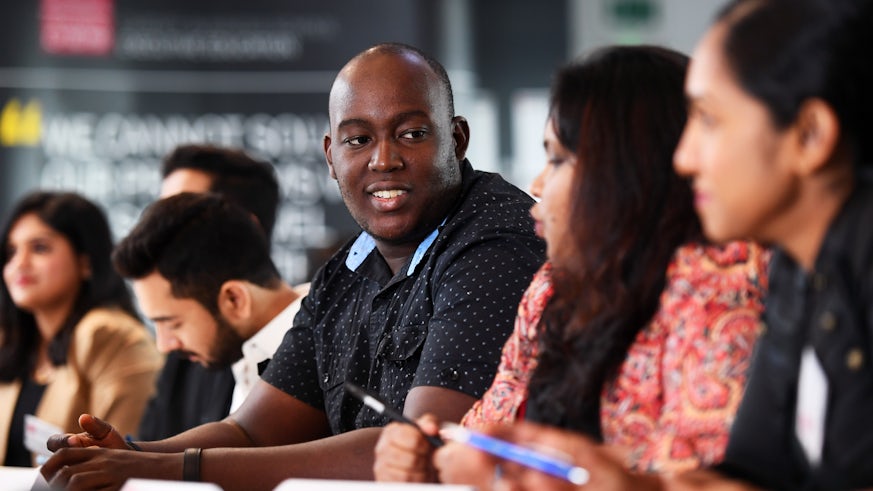 Students sitting in a row. The camera is focussing on a male student listening to a female student sat next to him talking.