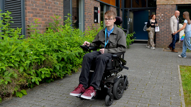 A person in a motorised wheelchair on a pathway near a brick building, with others walking nearby.
