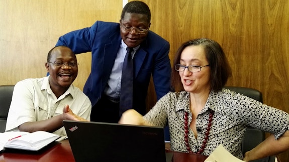 Dr Nelson Mlambo sitting with Professor Loredana Polezzi, looking at a laptop, with Professor Jairos Kangira standing behind