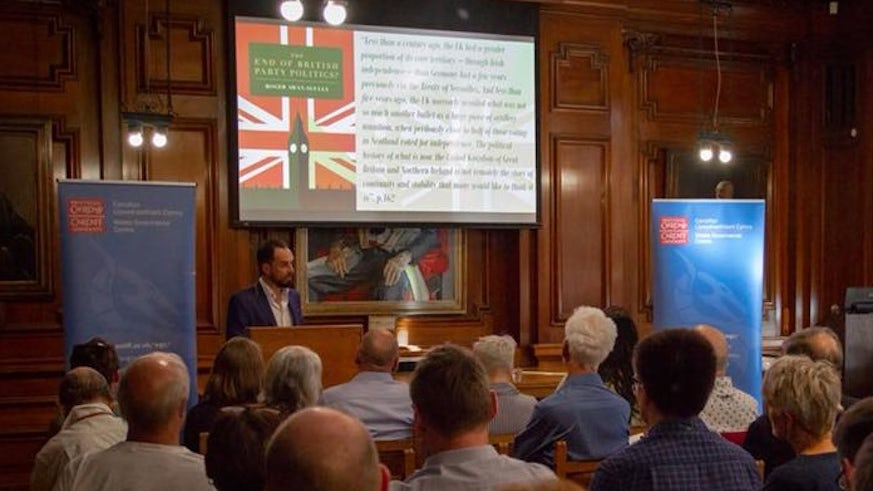Professor Roger Awan-Scully stands speaks in front of a busy room at a launch event. 