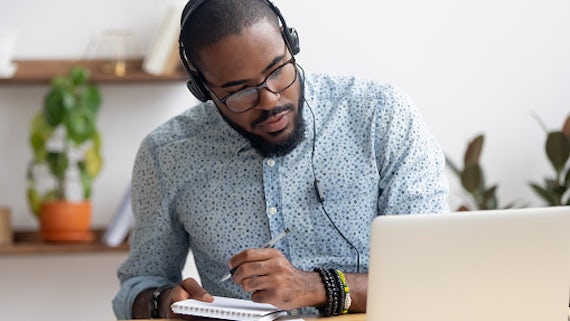 a Black man wearing headphones sits at a laptop making notes