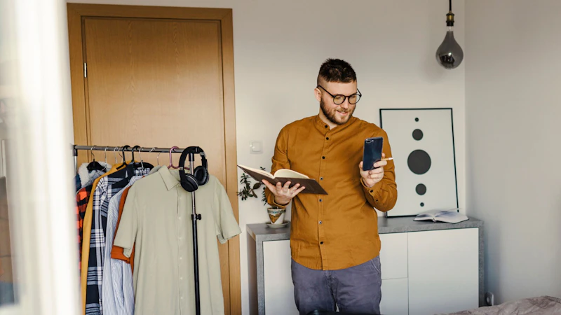 Male standing in his bedroom packing a suitcase. He's checking his phone whilst also holding a notebook. 