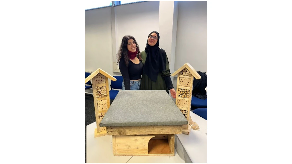 An image of two women standing indoors with two insect hotels and a hedgehog house