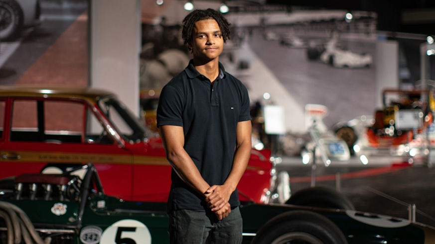 A portrait of a young black man wearing a black polo shirt. Behind him and out of focus are classic red racing cars.