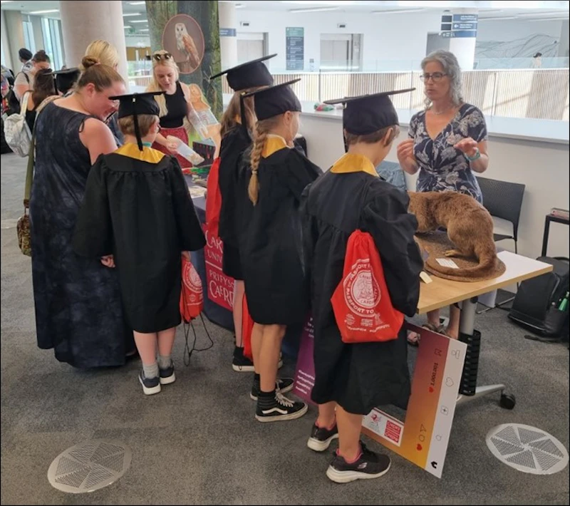 A group of children wearing graduation caps and gowns standing around a table speaking to Dr Liz Chadwick about otters. There is a taxidermy otter on the table.