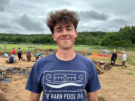 A close-up shot of a man smiling with an archaeological dig-site in the background.