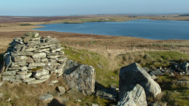 The flat stones and large rocks of the cairn in the green landscape facing the still water of the loch. 