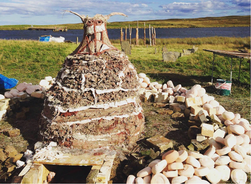 A kiln shaped like a mound that has sculture features at the top including a geometric pattern and two short sticks that extend from each side at the top in a 90 degrees angle. The kiln sits in a circle of smooth round pale coloured stones. 