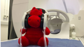 The mascot Dylan the red dragon sat on top of the bed of the MRI scanner wearing headphones