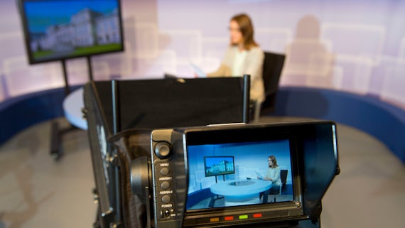 News camera pointing at a woman sitting at a table next to a screen.