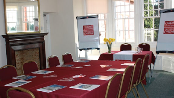 Room with table and chairs, laid with red tablecloth, papers on table, a table at front of room and flipcharts.