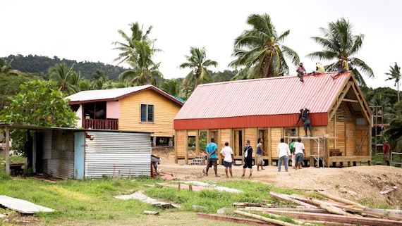 A group of builders work on a wood building - there are palm trees in the background